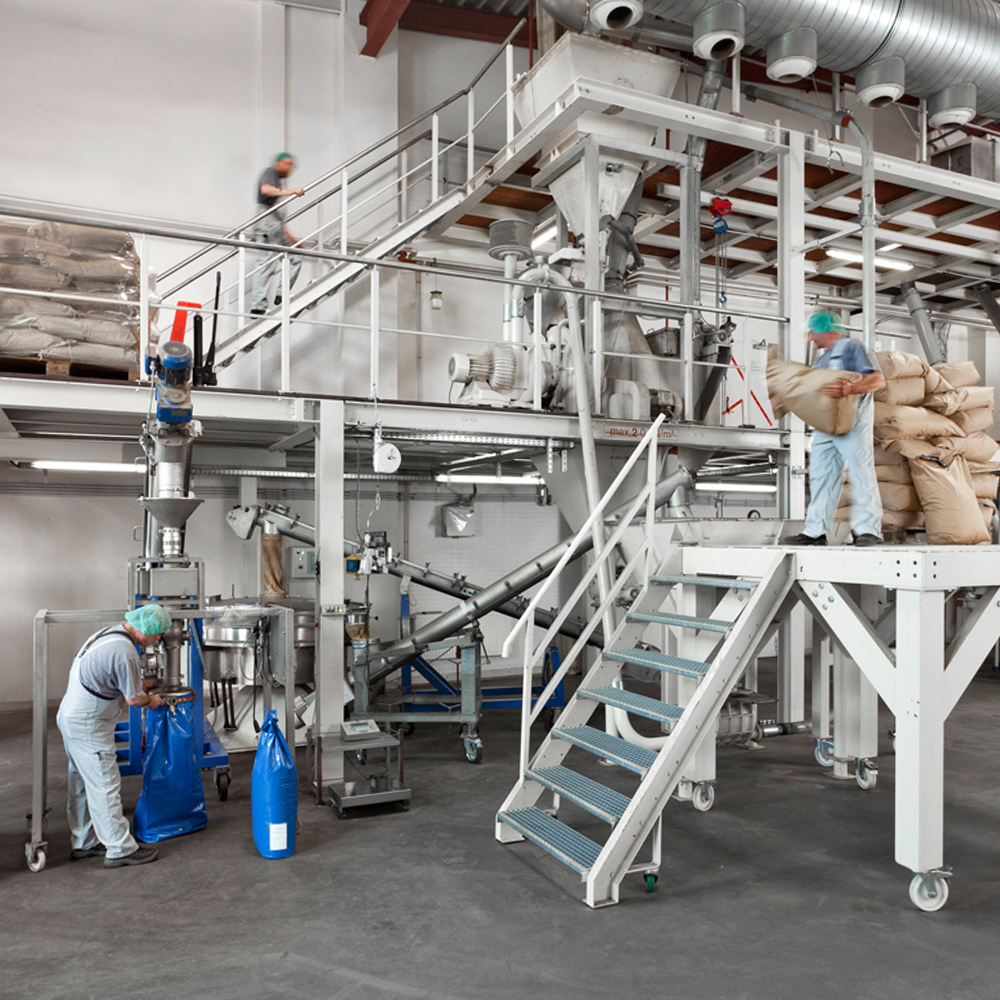 In einer Fabrik sind Arbeiter mit Schutzhelm und -kleidung zu sehen. Einer ist auf einer Treppe und trägt einen Sack, während ein anderer am Maschinenbereich mit blauen Säcken arbeitet. Im Hintergrund sind Maschinen und Förderbänder zu erkennen.