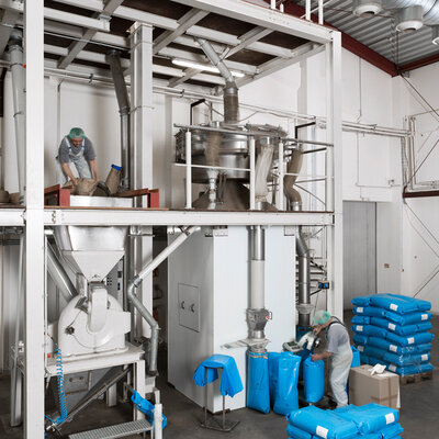 In einer Produktionshalle sind zwei Arbeiter beschäftigt. Einer steht auf einer Plattform und befüllt Säcke, während der andere am Boden blaue Säcke mit einem Gerät verschließt. Im Hintergrund sind Maschinen und Förderanlagen zu sehen.
