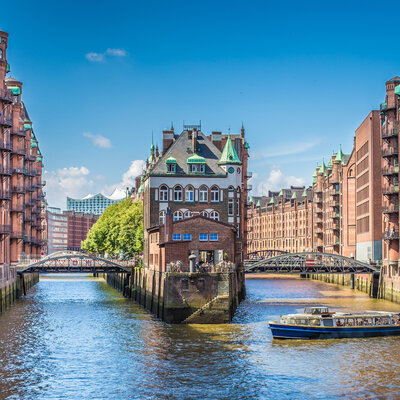 Ein malerischer Blick auf den Hafen mit einer historischen Backsteinarchitektur. Im Vordergrund fließt ein schmaler Wasserkanal, an dem ein Ausflugsboot fährt. An den Ufern stehen alte Gebäude, teilweise mit grünen Dächern, unter einem blauen Himmel mit wenigen Wolken.
